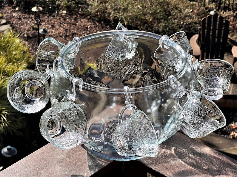 May include: A clear glass punch bowl with a set of matching cups hanging from the rim. The cups have a textured design and are perfect for serving punch at a party.