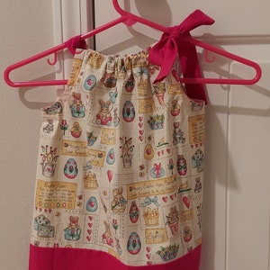 May include: A pink and white sleeveless dress with a bow at the neck. The dress has a pattern of bunnies, eggs, and flowers with the text "Happy Easter Happy Spring" and "Look forward to the joy they bring".
