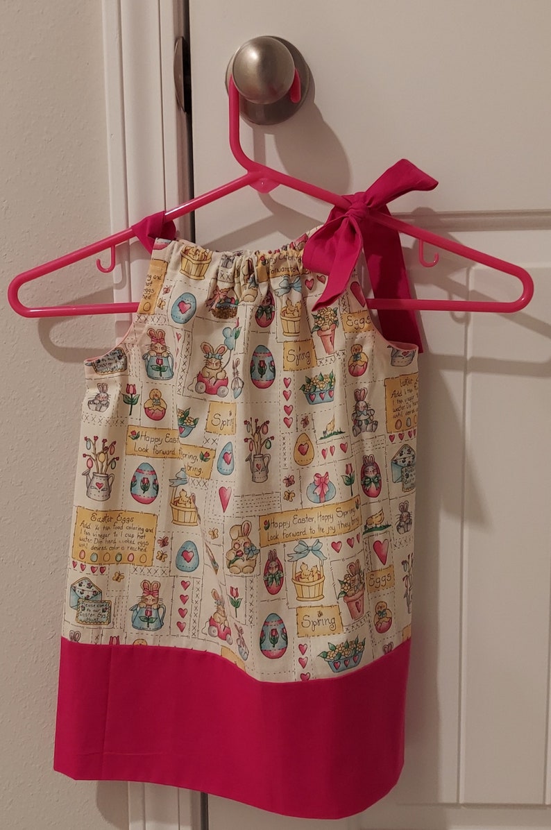 May include: A pink and white sleeveless dress with a bow at the neck. The dress has a pattern of bunnies, eggs, and flowers with the text "Happy Easter Happy Spring" and "Look forward to the joy they bring".