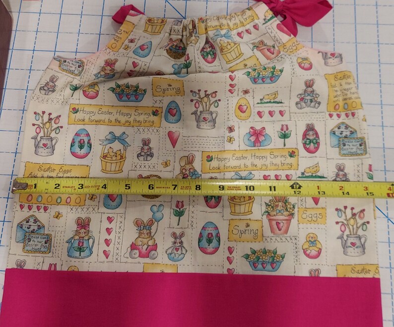 May include: A pink and white fabric with a repeating pattern of Easter eggs, bunnies, chicks, and flowers. The fabric has the text "Happy Easter, Happy Spring" and "Look forward to the joy they bring".