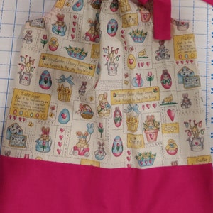 May include: A sleeveless, white cotton dress with a pink trim. The dress has a pattern of bunnies, eggs, flowers, and the words "Happy Easter, Happy Spring, Look forward to the joy they bring." The dress has a pink bow at the top.