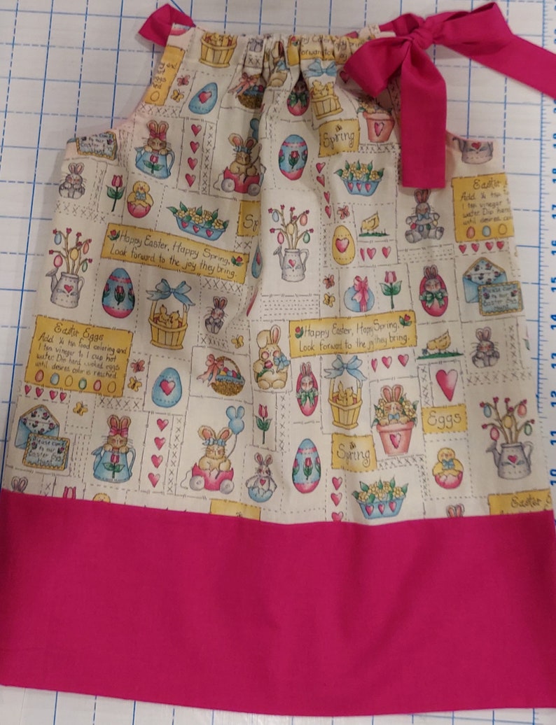May include: A sleeveless, white cotton dress with a pink trim. The dress has a pattern of bunnies, eggs, flowers, and the words "Happy Easter, Happy Spring, Look forward to the joy they bring." The dress has a pink bow at the top.