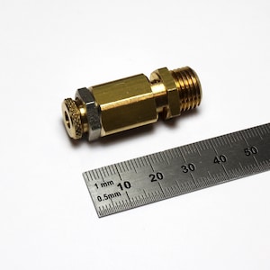 Puede incluir: Un racor neumático de latón con un extremo roscado y una tuerca moleteada. Una regla metálica con marcas de milímetros está junto al racor. El racor mide aproximadamente 5 cm de largo.