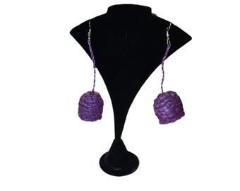 May include: A pair of purple woven earrings with a spherical design. The earrings hang from silver-colored hooks and are displayed on a black jewelry stand against a white background. The earrings have a textured appearance.
