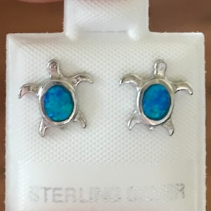 May include: A pair of sterling silver stud earrings shaped like sea turtles. Each earring features a central oval-shaped blue opal inlay within the silver frame. The earrings are attached to a white display card with the text STERLING SILVER.