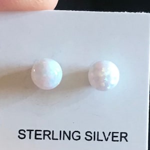 May include: A pair of round, white opal stud earrings. The earrings are set on a white card with the words "STERLING SILVER" printed in black. The opals have a subtle iridescent shimmer.