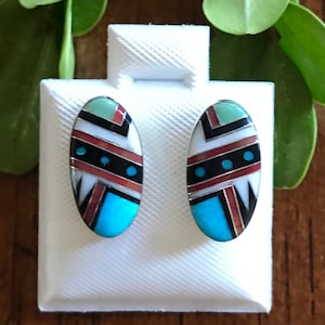 May include: A pair of oval-shaped earrings with a geometric design. The earrings are made of silver and feature turquoise, red, black, white, and green stones.