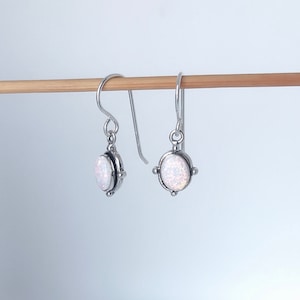Pode incluir: Um par de brincos pendurados prateados. Cada brinco apresenta uma pedra oval branca iridescente emoldurada por um design decorativo. Os brincos pendem de um gancho simples, um estilo de joalharia clássico.