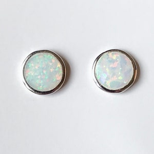 May include: Two round stud earrings with a white opal center and a silver-colored bezel. The opals have a shimmering, iridescent quality, reflecting various colors. The earrings are set against a plain white background.