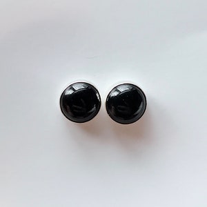 May include: Two round, black stud earrings with a glossy finish. The earrings are set in a silver-colored metal. The simple design makes them suitable for everyday wear.