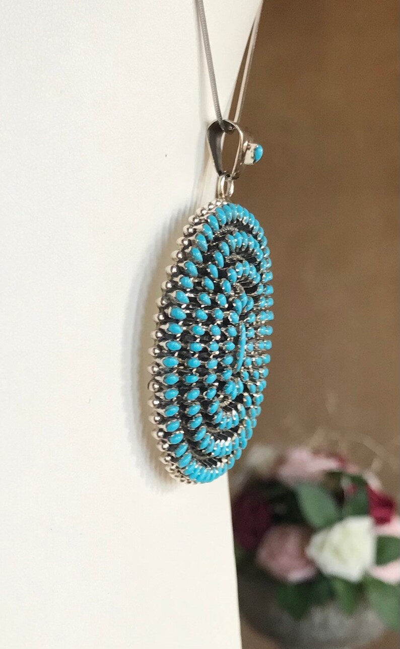 Pu&ograve; includere: Una collana con un ciondolo in turchese e argento. Il ciondolo &egrave; rotondo, piatto e ha un'intarsio in turchese. Il ciondolo &egrave; circondato da un bordo in argento.