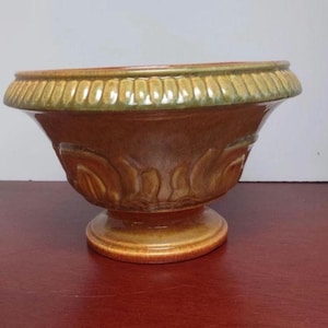 Puede incluir: Una urna decorativa de cerámica con un borde ancho acanalado y una base de pedestal. La urna presenta un color marrón cálido con detalles en verde y dorado. El cuerpo de la urna tiene un diseño en relieve detallado. Adecuado para la decoración del hogar.