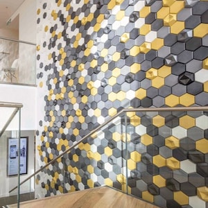 Könnte beinhalten: Eine Wand, die mit einem geometrischen Muster aus sechseckigen Fliesen in Grautönen, Weiß und Gold bedeckt ist. Die Fliesen haben einen 3D-Effekt und erzeugen eine strukturierte Oberfläche.