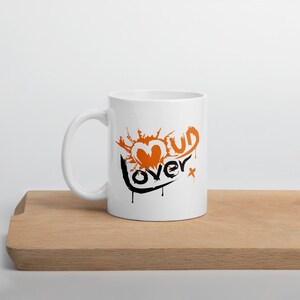 May include: White ceramic mug with a black and orange graphic that says "Mud Lover" with a heart shape in the middle.