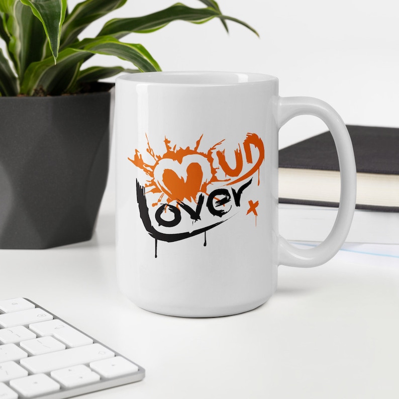 May include: White ceramic mug with a black and orange graphic that says "Mud Lover" with a heart shape and splattered paint design.