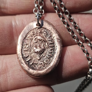 May include: An oval-shaped copper pendant with a textured, stamped surface featuring a central emblem and surrounding lettering, suspended from a silver-toned metal chain link necklace. The pendant has a rustic, metallic finish with visible details.