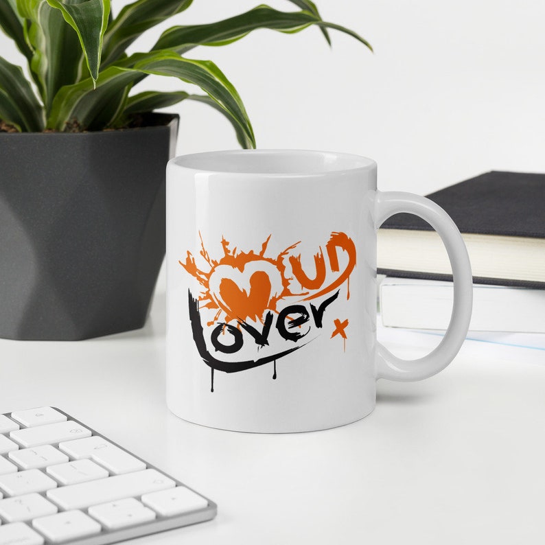 May include: White ceramic mug with a black and orange graphic that says "Mud Lover" with a heart shape in the middle.