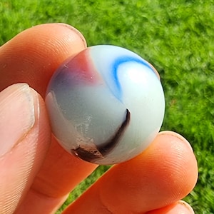Może przedstawiać: Biały szklany kulkowy z niebieskimi, czerwonymi i brązowymi smugami. Kulka jest trzymana między dwoma palcami.