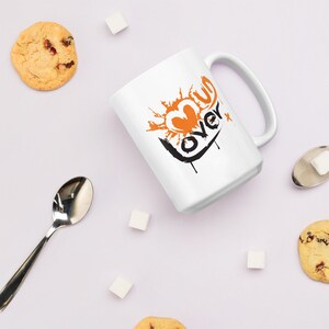May include: White ceramic mug with a black and orange graphic design that says "Love Over" with a heart shape. The mug is on a white surface with sugar cubes and cookies.