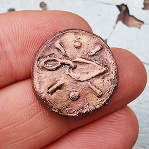 May include: A copper coin with a design of a stylized bird with its wings spread. The coin has a raised rim and a smooth surface.