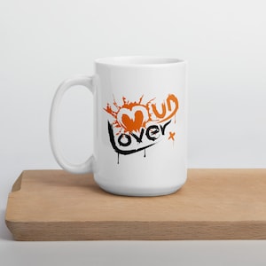 May include: White ceramic mug with a black and orange graphic that says "Mud Lover" with a heart shape in the middle.