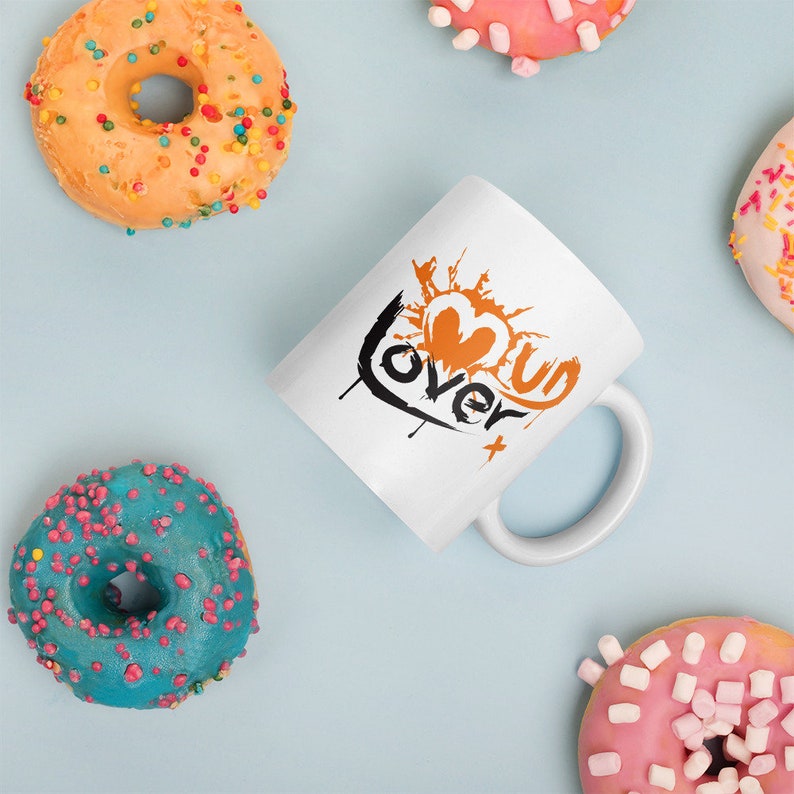 May include: White ceramic mug with a black and orange graphic design. The graphic features a heart with the word "over" written around it. The word "us" is written inside the heart.