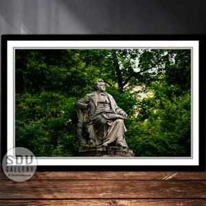 May include: A framed photograph of a stone statue of a seated man, set against a backdrop of lush green trees. The statue is detailed, with the man appearing to be reading or writing. The photograph is in a black frame, with the SDU Gallery logo in the lower left corner.