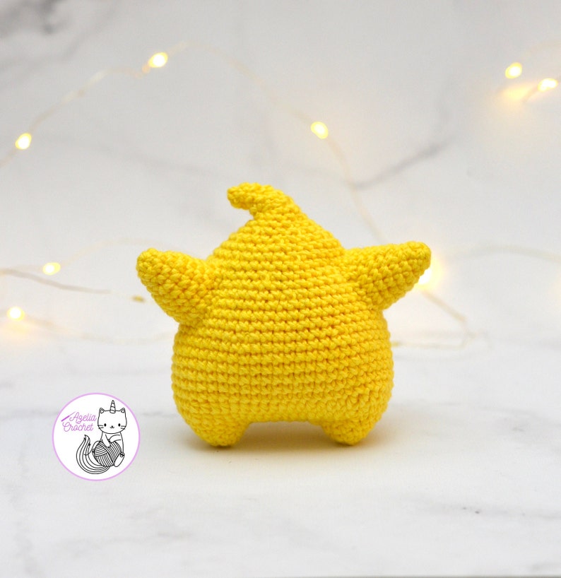 Puede incluir: Un juguete de peluche de color amarillo en forma de estrella tejido a crochet. El juguete est&aacute; sobre una superficie de m&aacute;rmol blanco.