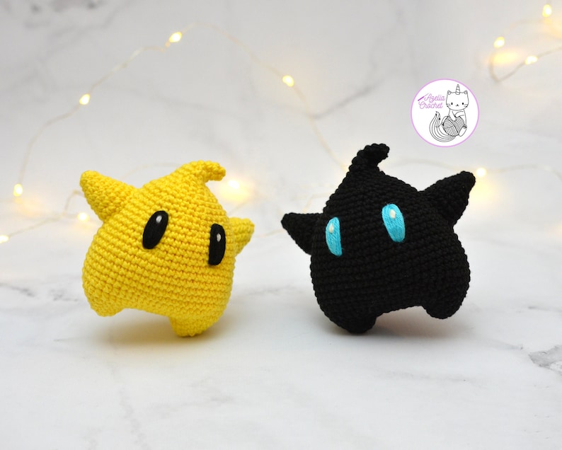 Puede incluir: Dos peluches de crochet, uno amarillo y otro negro, con forma de estrellas y ojos grandes. El peluche amarillo tiene ojos negros y el peluche negro tiene ojos azules.