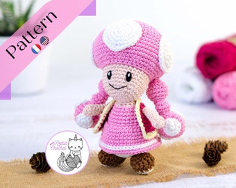 May include: Pink crocheted mushroom character with white accents, wearing a pink dress and a yellow sash. The character has a smiling face with black eyes and a brown nose. The character is standing on a brown surface with pine cones.