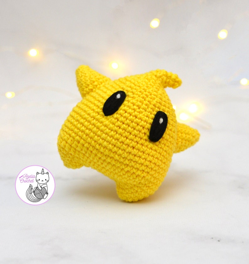 Puede incluir: Un juguete de peluche amarillo de crochet con forma de fantasma con ojos negros. El juguete est&aacute; hecho con un hilo suave y texturizado y tiene una forma redondeada y bulbosa.