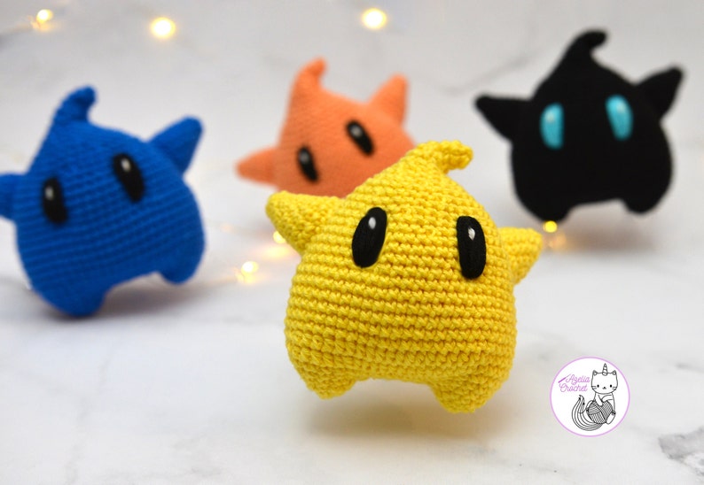 Puede incluir: Cuatro peluches de estrella de ganchillo en diferentes colores: azul, naranja, amarillo y negro. Los peluches tienen ojos negros y est&aacute;n sentados sobre una superficie blanca.