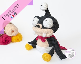 May include: Crochet pattern for a black and white alien character with a red cape. The pattern is available in English and French. The image shows the finished crochet character sitting on a white surface.