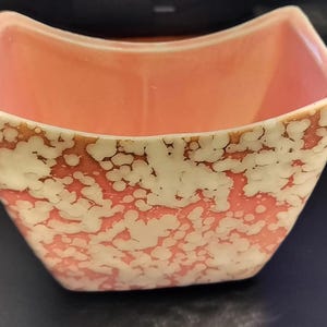 May include: A square ceramic bowl with a peach-colored interior and a speckled exterior design in white and coral. The rim has a brown trim. This decorative bowl is ideal for serving snacks or as a decorative piece.