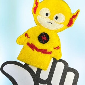 May include: A yellow felt Flash superhero finger puppet with red lightning bolt accents and a black lightning bolt emblem. The puppet is on a white foam hand with a black outline, against a light blue background.