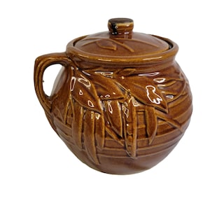 Puede incluir: Una olla de cerámica marrón con tapa, decorada con un diseño de hojas. La olla tiene un asa y es perfecta para guardar objetos pequeños.