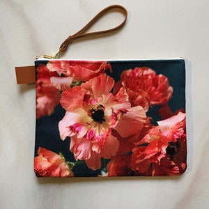 May include: A black canvas zippered pouch with a floral print. The print features pink and red flowers on a dark background. The pouch has a brown leather strap.
