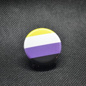 Puede incluir: Un botón redondo con los colores de la bandera del orgullo no binario. El botón tiene rayas amarillas, blancas, moradas y negras.