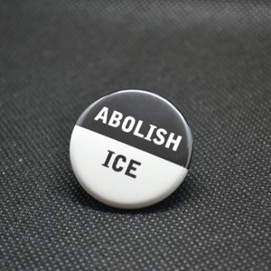 May include: A black and white button with the text "ABOLISH ICE" printed on it.