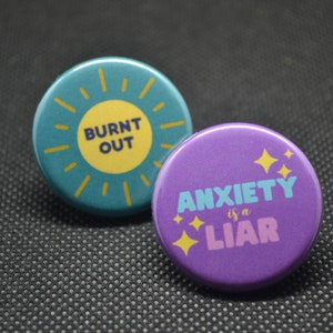 May include: Two round button pins with motivational messages. The first pin is teal blue with a yellow sun and the text "BURNT OUT". The second pin is purple with gold stars and the text "ANXIETY is a LIAR".