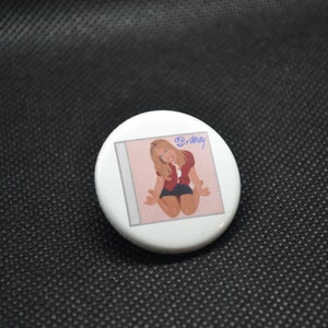 May include: A white button with a pink and white album cover featuring a woman in a red shirt and black skirt. The album cover has the text "Britney" in blue.