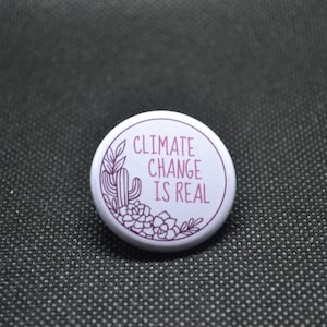 May include: White button with a pink outline and a cactus and succulent design. The text "Climate Change Is Real" is printed in pink.