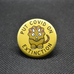 May include: Yellow button with the text "PUT COVID ON EXTINCTION" and a cartoon illustration of a brown and yellow dinosaur with a red and brown frill.