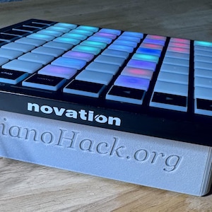 Może przedstawiać: Czarny kontroler MIDI Novation z siatką kwadratowych, podświetlanych przycisków w kolorze białym, niebieskim i czerwonym. Urządzenie jest uniesione na jasnoszarym stojaku z wytłoczonym napisem PianoHack.org z przodu, z podłączonym kablem USB.