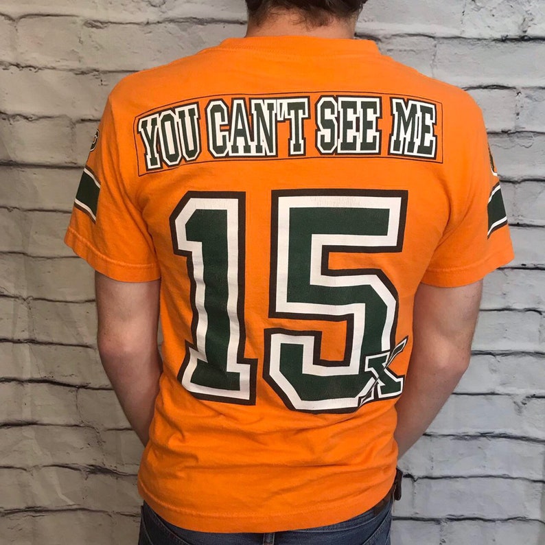 Puede incluir: Camiseta naranja con ribetes verdes y blancos. La parte posterior de la camiseta tiene el n&uacute;mero "15" y el texto "YOU CAN'T SEE ME".