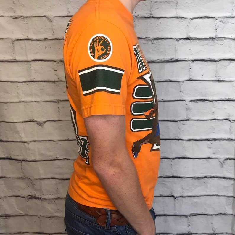 Puede incluir: Camiseta naranja con un gr&aacute;fico verde y blanco de un jugador de baloncesto. La camiseta tiene una franja verde y blanca en la manga y un parche circular con las palabras "Hustle Loyalty Respect" en el pecho izquierdo.