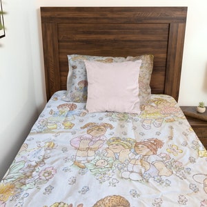 Pode incluir: Uma cama de solteiro com cabeceira de madeira, com um edredom estampado em azul claro e branco com personagens de desenhos animados. Uma almofada rosa claro repousa sobre a cama. O quarto tem uma mesa de cabeceira com uma lâmpada.