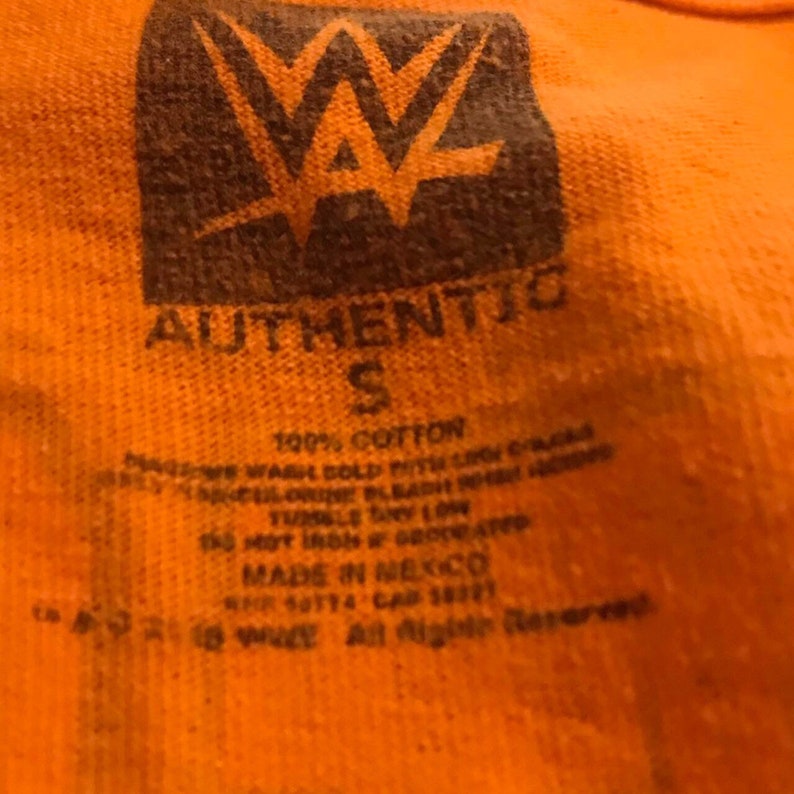 Puede incluir: Un primer plano de una etiqueta de camiseta naranja con las palabras "AUTHENTIC" y "S" impresas en ella. La etiqueta tambi&eacute;n incluye instrucciones de cuidado y el texto "100% ALGOD&Oacute;N", "HECHO EN M&Eacute;XICO" y "&copy; 2014 Todos los derechos reservados".
