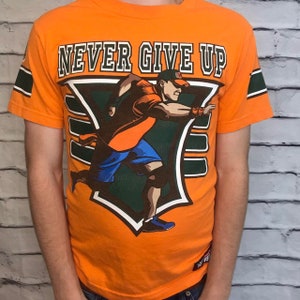 Puede incluir: Camiseta naranja con un gr&aacute;fico verde y blanco de un luchador corriendo. El gr&aacute;fico incluye el texto "NEVER GIVE UP".