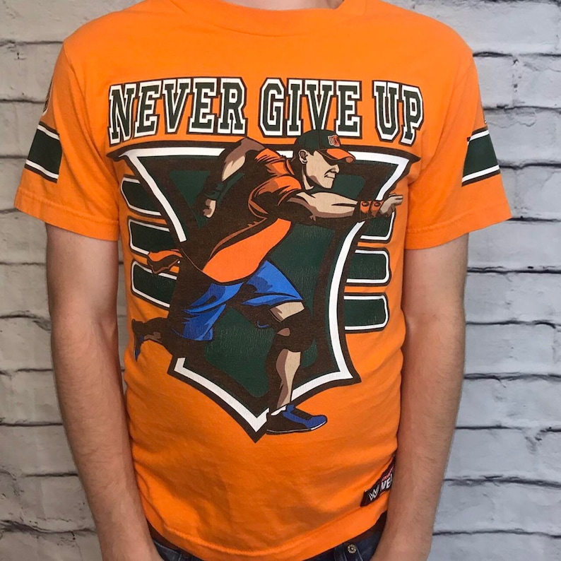 Puede incluir: Camiseta naranja con un gr&aacute;fico verde y blanco de un luchador corriendo. El gr&aacute;fico incluye el texto "NEVER GIVE UP".